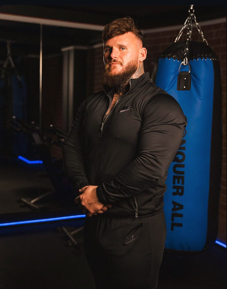Man in a gym standing in front of a blue punching bag, wearing a black athletic outfit, with arms crossed.